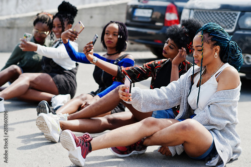 Group of five african american woman sitting on road together against suv car on parking and making selfie on phone.