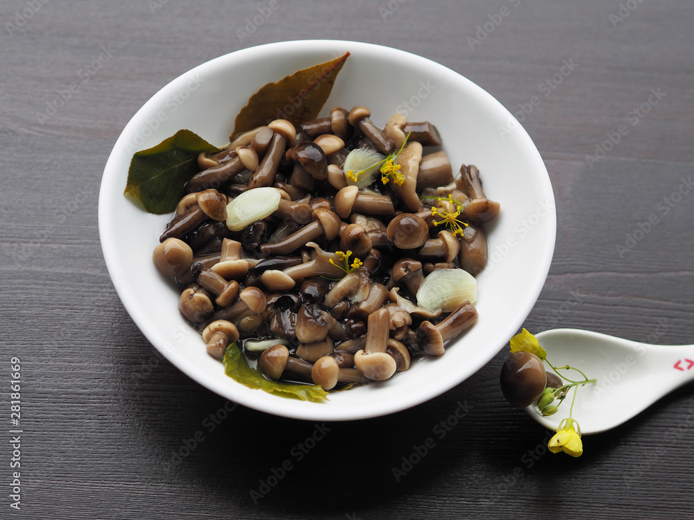Delicious and beautiful pickled mushrooms and spices in a white plate, one fungus in a spoon on a wooden background. Home recipe pickles - small mushrooms with garlic and herbs close-up. Delicious and beautiful pickled mushrooms and spices in a white plate, one fungus in a spoon on a wooden background. Home recipe pickles - small mushrooms with garlic and herbs close-up.