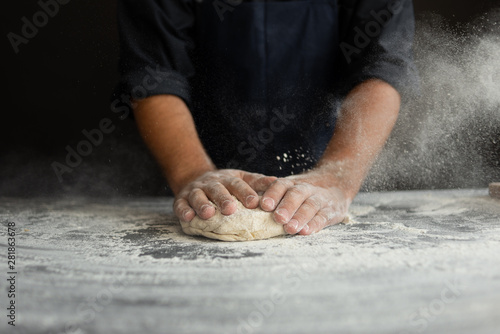 chef kneads raw dough, dust from flour flies in different directions, male hands on a black background