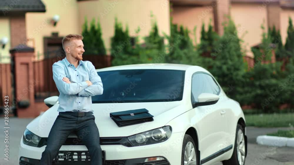 Happy successful young business male making winner gesture rising up hand sitting on car bonnet Happy successful young business male making winner gesture rising up hand sitting on car bonnet