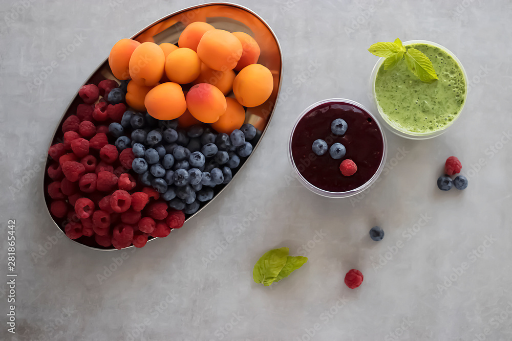 Fruit and berry smoothies and green smoothie. Fresh apricots, raspberries and blueberries on an oval metal plate. Gray background. Top view. Fruit and berry smoothies and green smoothie. Fresh apricots, raspberries and blueberries on an oval metal plate. Gray background. Top view.