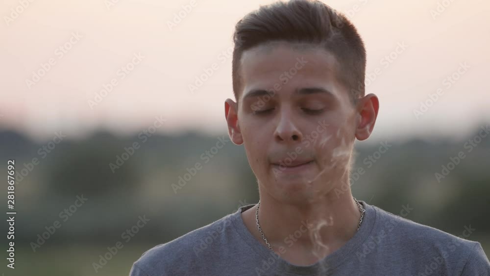 Closeup portrait of handsome stylish young boy with long brunette hair vaping electronic cigarette outdoors at sunset in hot summer day at sunset Closeup portrait of handsome stylish young boy with long brunette hair vaping electronic cigarette outdoors at sunset in hot summer day at sunset