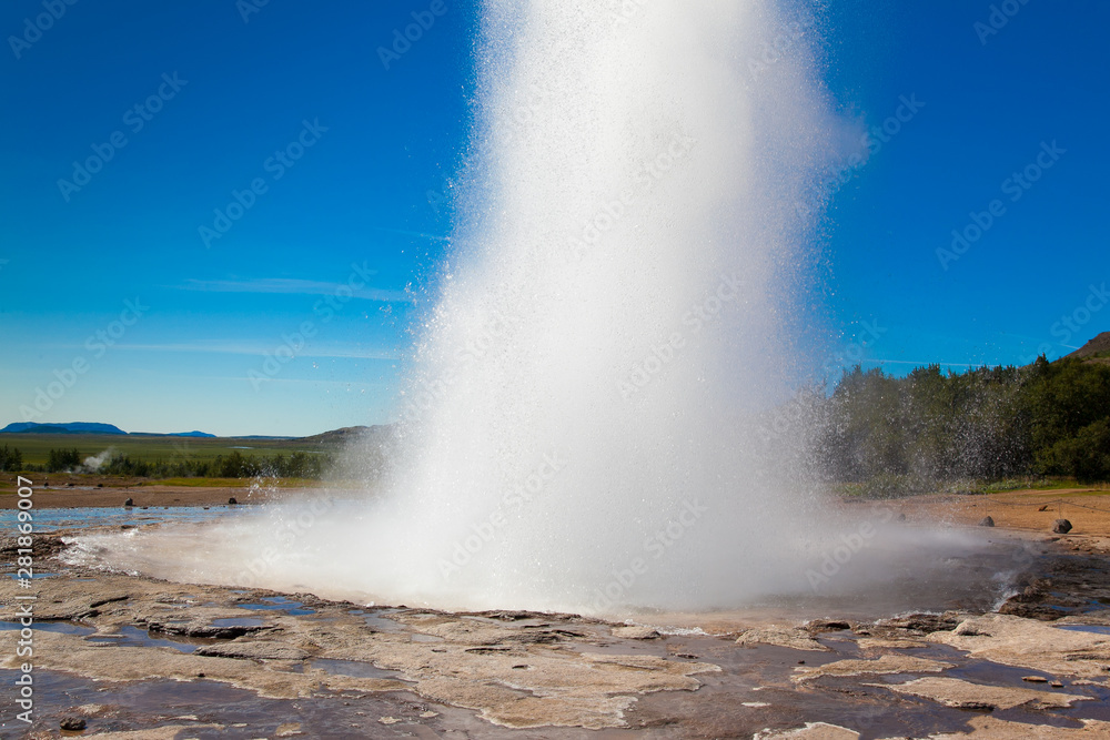 Geyser in Iceland against the blue sky and forest. A tall column of hot water breaks out of the ground. Nobody. Geyser in Iceland against the blue sky and forest. A tall column of hot water breaks out of the ground. Nobody.