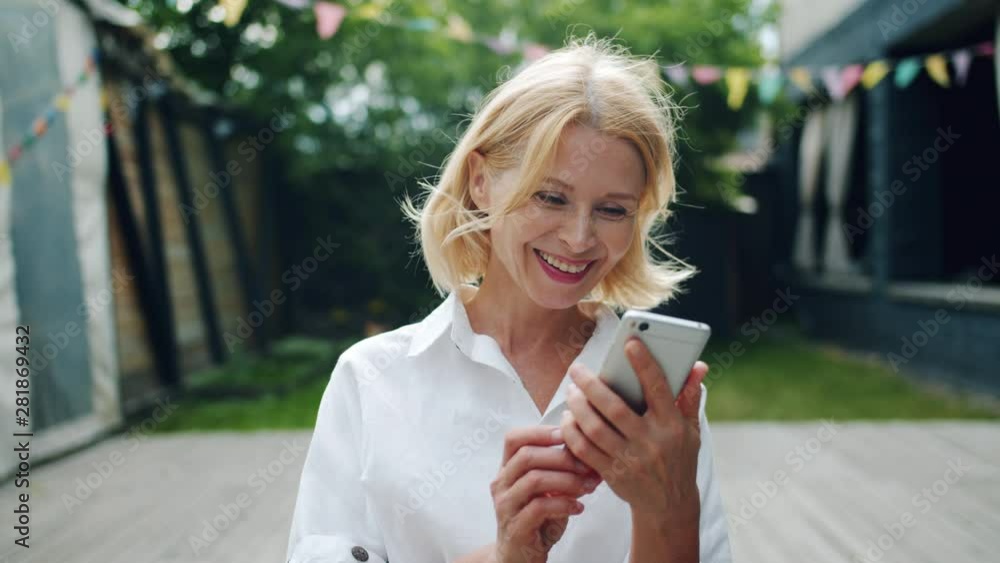 Joyful mature woman is taking smartphone looking at screen smiling enjoying good news standing outdoors in city street. Modern technology and emotions concept. Joyful mature woman is taking smartphone looking at screen smiling enjoying good news standing outdoors in city street. Modern technology and emotions concept.