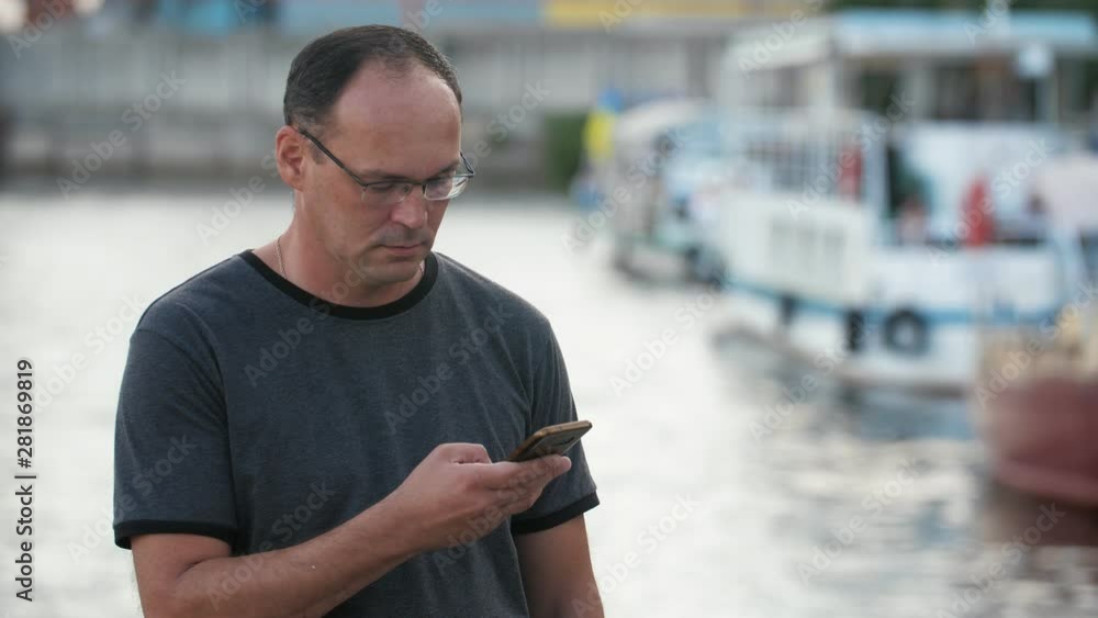 Calm adult man using smartphone on near the water on the embankment in the evening Calm adult man using smartphone on near the water on the embankment in the evening