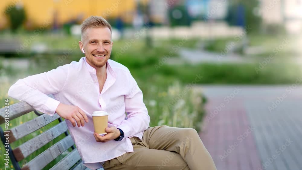 Happy casual guy in wireless earphones sitting on bench at summer park holding paper coffee cup Happy casual guy in wireless earphones sitting on bench at summer park holding paper coffee cup