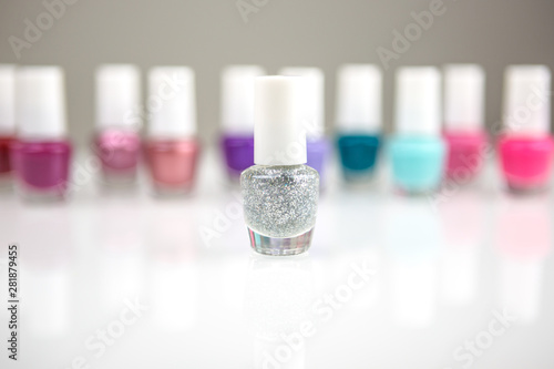 View on a shiny white desk full of small glass and plastic bottles with assorted nail polish and lacquer on a back creating row with focus on one white shiny sparling nail polish.