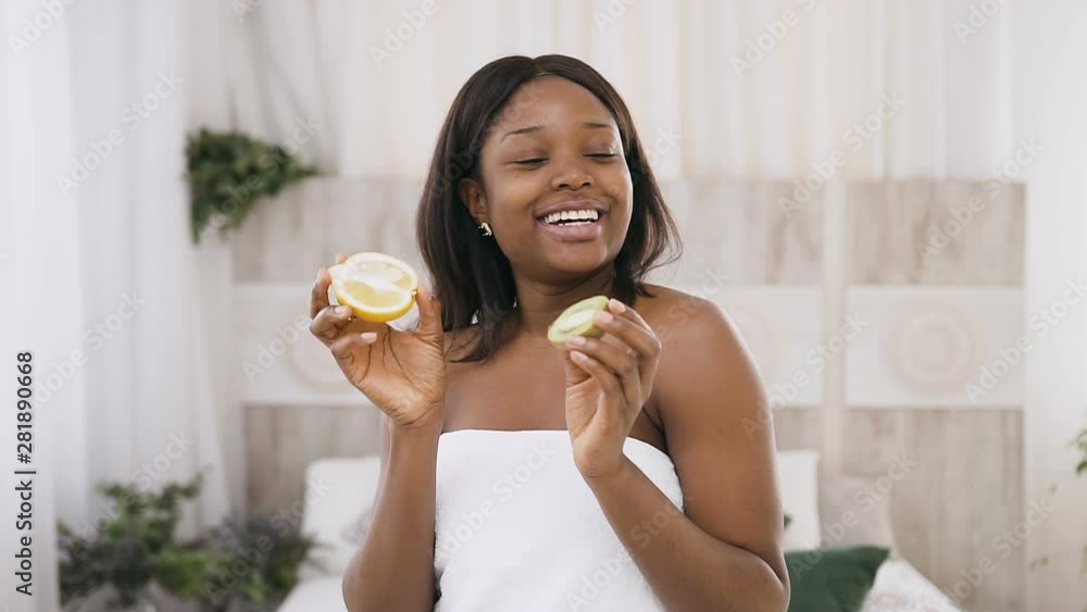 Portrait shot of funny amusing african american young woman holding kiwi and orange halves in hands over light background in spa. Healthy skin care. Healthy lifestyle concept Portrait shot of funny amusing african american young woman holding kiwi and orange halves in hands over light background in spa. Healthy skin care. Healthy lifestyle concept