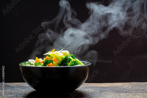 Fototapeta Naklejka Na Ścianę i Meble -  The steam from the vegetables carrot broccoli Cauliflower in a black bowl , a steaming. Boiled hot Healthy food on table on black background,hot food and healthy meal concept