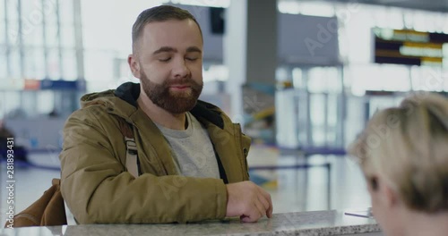 Close up of the young Caucasian smiled guy with beard coming to the checking desk at the airport, giving his passport and ticket.
