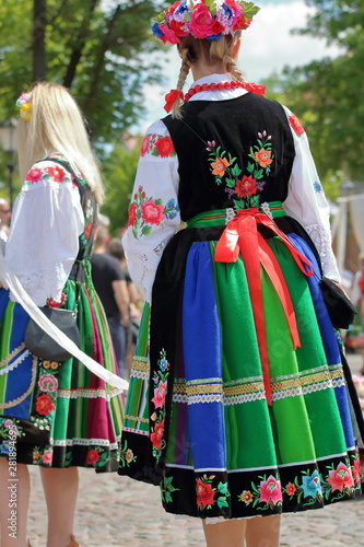 Young Polish girl stands on her back dressed in traditional folk female dress from lowicz region, in background another blond hair woman in folk costume and other people in soft focus, Corpus Christi 