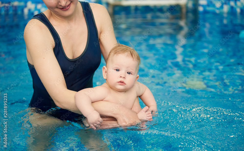 Cropped snapshot of young smiling mother holding her son sitting in her arms in pool in blue water. Baby looking in camera. Infant swimming and doing water exercises. Health promotion concept Cropped snapshot of young smiling mother holding her son sitting in her arms in pool in blue water. Baby looking in camera. Infant swimming and doing water exercises. Health promotion concept
