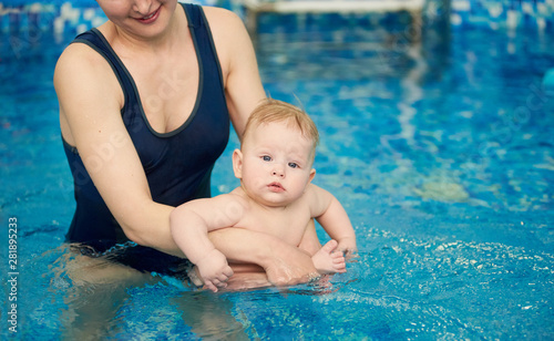 Cropped snapshot of young smiling mother holding her son sitting in her arms in pool in blue water. Baby looking in camera. Infant swimming and doing water exercises. Health promotion concept
