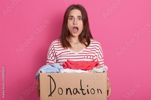 Image of shocked emotional lady with widely opened mouth and eyes, being surprised, having carton box with donated clothes in hands, looking directly at camera. People and volunteering concept. photo