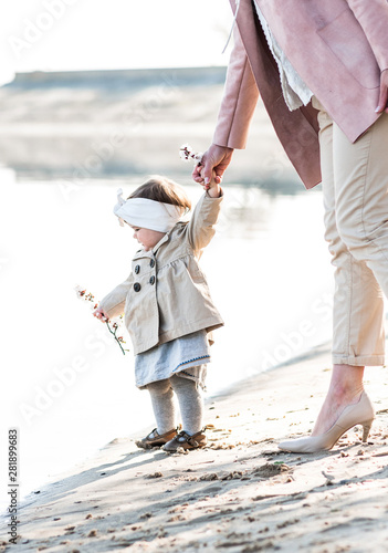 charming stylish baby girl holding mom's hand on the sandy shore near the river