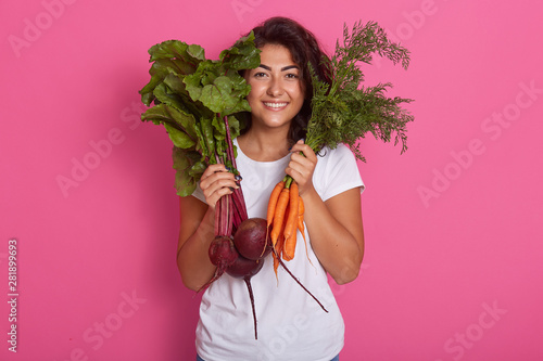 Close up portrait of attractive caucasian smiling woman isolated on rose studio background, wearing white casual t shirt, holding beets and carrots in hands. Raw dood eatinf and healthy eating concept photo