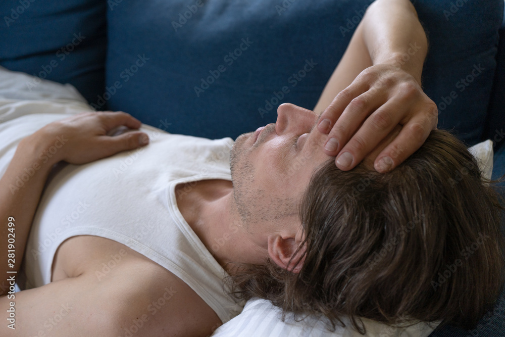 A man in a home sports suit lies at home on the sofa and put his hand on his head and another one on his stomach. Headache, stomachache, malaise, colds, fever. Side view A man in a home sports suit lies at home on the sofa and put his hand on his head and another one on his stomach. Headache, stomachache, malaise, colds, fever. Side view