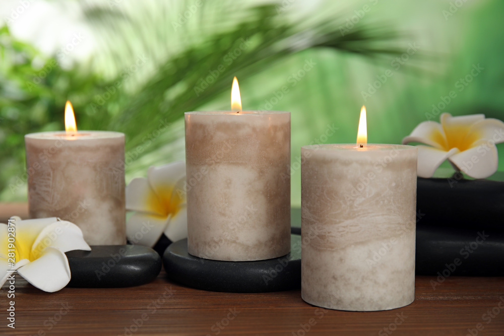 Burning candles and plumeria flowers on wooden table against blurred green background