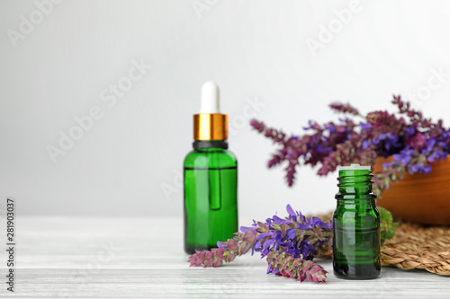 Bottles of sage essential oil and flowers on white table  space for text