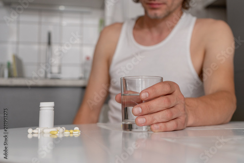 A man sits at the white kitchen table, holds a glass of water, going to drink pills and tablets lying on the table. Illness, malaise, cold, ache