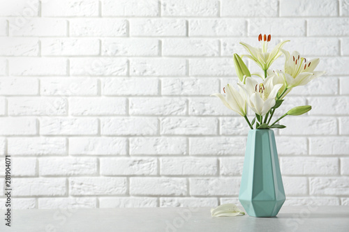 Vase of beautiful lilies on table against  white brick wall, space for text