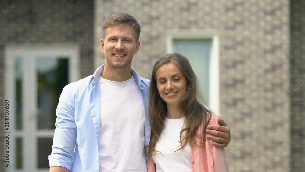 Joyful young couple near for sale sign smiling camera standing outside house Joyful young couple near for sale sign smiling camera standing outside house