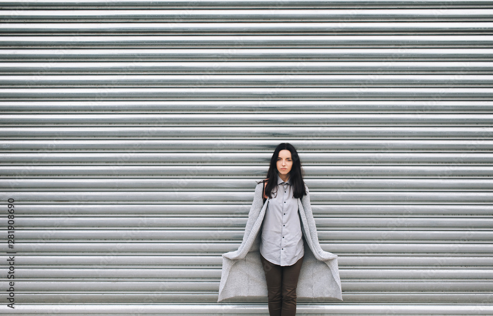 A young European girl in a gray cardigan stands against a metal textured roller shutters, looking skeptical and holds with her hands in pockets. Casual clothes. Copy space, background. A young European girl in a gray cardigan stands against a metal textured roller shutters, looking skeptical and holds with her hands in pockets. Casual clothes. Copy space, background.