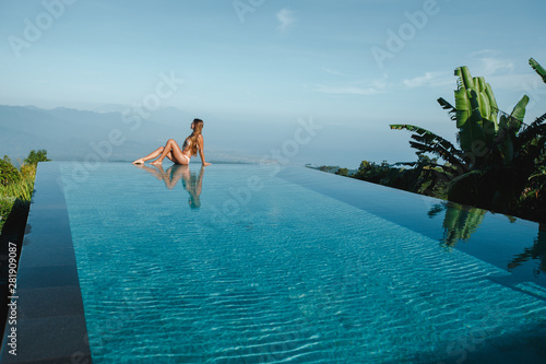 Slim young woman in swimsuit relaxing on edge tropical infinity pool in mountains. Palms around and crystal clean water. Luxury resort on Bali island