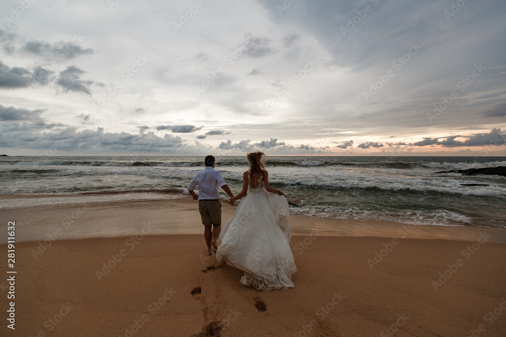 Young beautiful newlyweds in love holding hands and walking away on tropical sandy beach, back view Young beautiful newlyweds in love holding hands and walking away on tropical sandy beach, back view