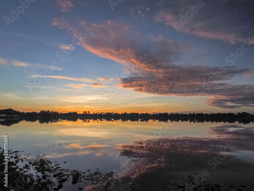 Panorama Lake view morning of fat clouds moving above reservoir and reflection in water with yellow sun light in the sky background, sunrise at Krajub reservoir, Ban Pong, Ratchaburi, Thailand. © Yuttana Joe