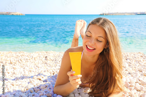 Happy smiling woman holding sun cream tube protection lying on pebbles beach. Sunscreen girl looking suntan lotion in plastic container.