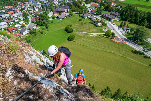 Women on a via ferrata route on Grancisce (Grancise) hill, above the Mojstrana village in Slovenia, on a sunny day, with tiny houses visible below. Outdoor adventures concept in Slovenia.