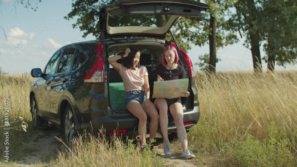 Multiethnic charming female friends with road map looking for directions while sitting in car trunk. Cheerful traveler women searching location with paper map during summer vacations road trip. Multiethnic charming female friends with road map looking for directions while sitting in car trunk. Cheerful traveler women searching location with paper map during summer vacations road trip.