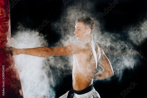 Handsome shirtless male boxer practicing on a punching bag indoors
