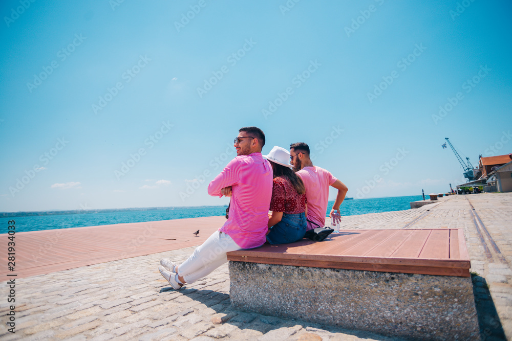 Four tourist friends having fun and taking pictures on a modern abstract bench paced on a seaside marine port on a sunny day. Enjoying the beautiful blue and turquoise sea.