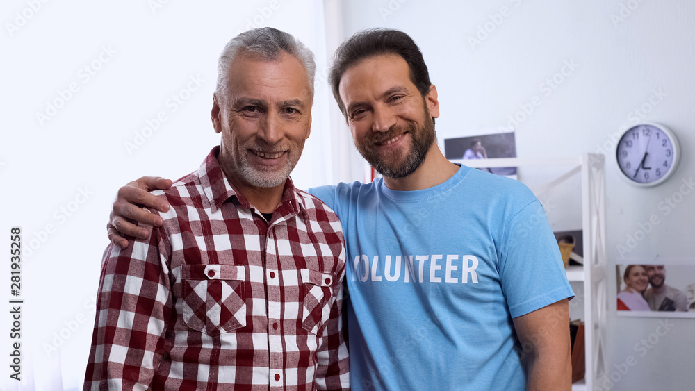 Young male volunteer hugging senior male and smiling at camera, charity concept Young male volunteer hugging senior male and smiling at camera, charity concept