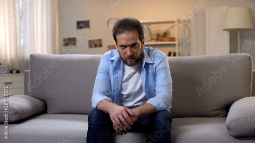 Sad lonely man aggressively looking into camera sitting on sofa at home, problem