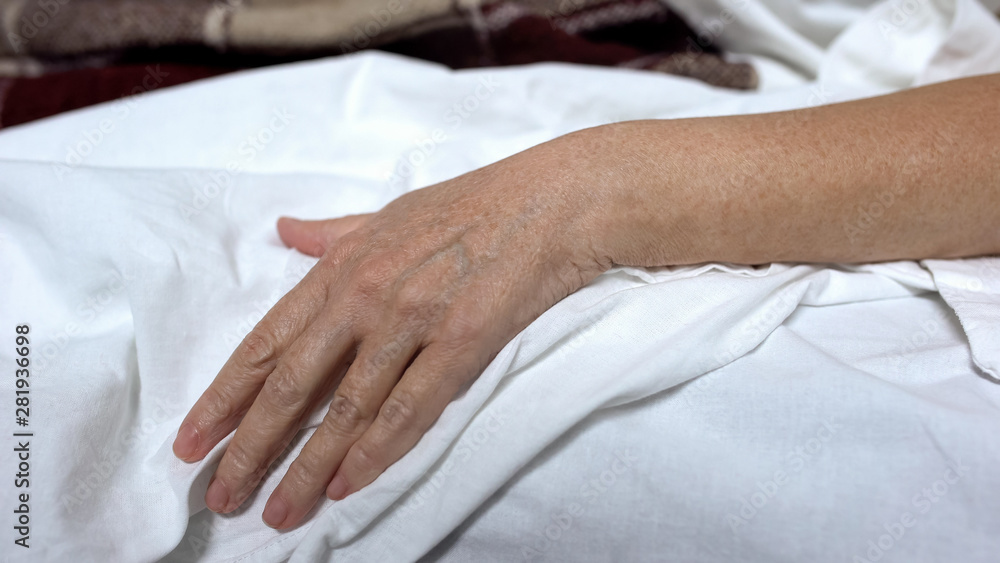 Senior woman hand on bed linen, lying in sick bed, suffering illness, death risk