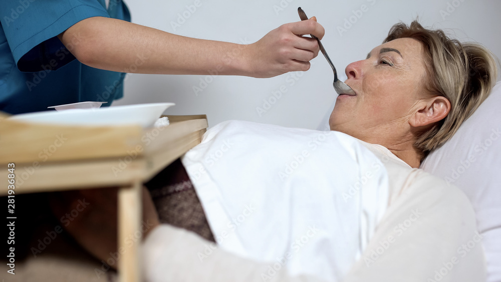Fototapeta premium Nurse feeding disabled mature woman with porridge, care for patients, hospice