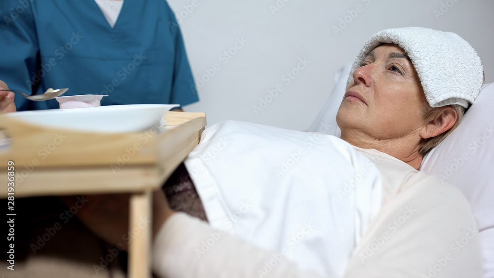 Obraz premium Nurse feeding sick indifferent woman with compress on head, lying in hospice bed