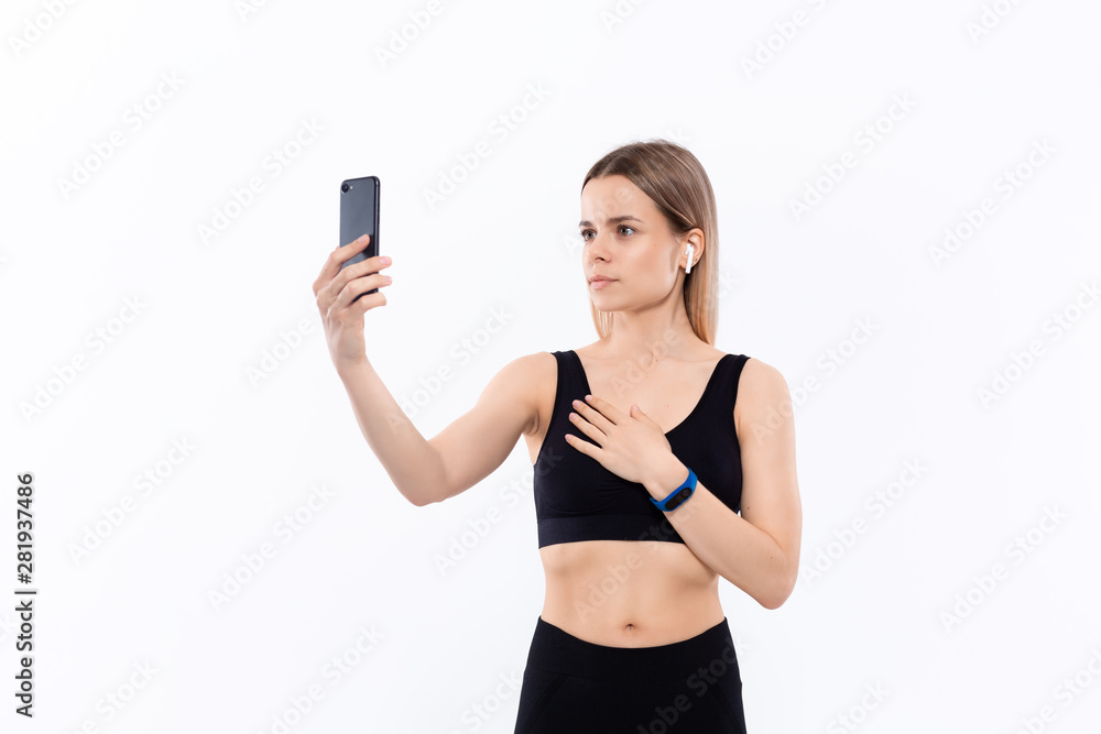 Young sporty blond woman in a black sportswear making selfie with smartphone showing bicep after workout standing over white background. Model listens music during workout with wireless headphones Young sporty blond woman in a black sportswear making selfie with smartphone showing bicep after workout standing over white background. Model listens music during workout with wireless headphones