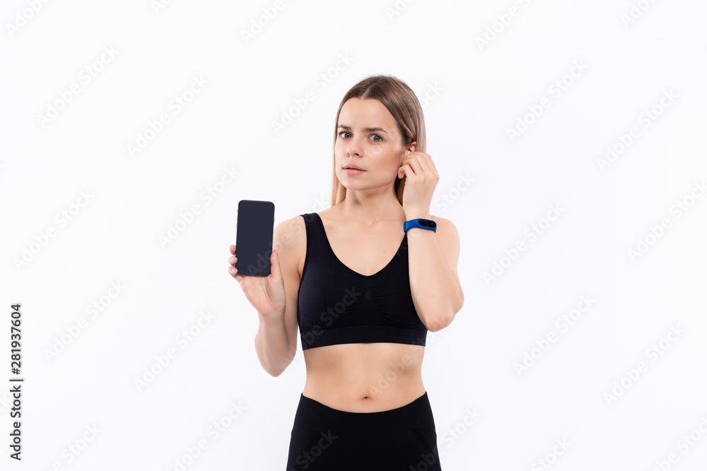 Young sporty blond woman in a black sportswear with wireless ear buds showing screen of a smartphone with a finger standing over white background. Young sporty blond woman in a black sportswear with wireless ear buds showing screen of a smartphone with a finger standing over white background.