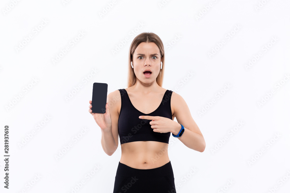 Surprised young sporty blond woman in a black sportswear with wireless ear buds showing screen of a smartphone with a finger standing over white background Surprised young sporty blond woman in a black sportswear with wireless ear buds showing screen of a smartphone with a finger standing over white background