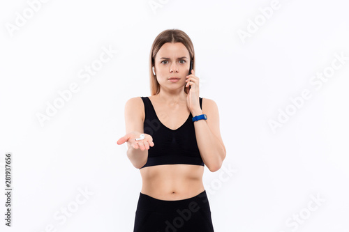 Astonished young sporty blond woman in a black sportswear with wireless ear buds speaking on the phone standing over white background.