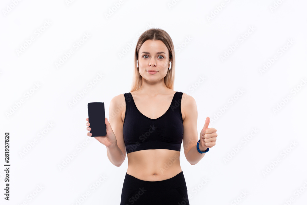 Excited young sporty blond woman in a black sportswear with wireless ear buds showing screen of a smartphone with a finger standing over white background. Excited young sporty blond woman in a black sportswear with wireless ear buds showing screen of a smartphone with a finger standing over white background.