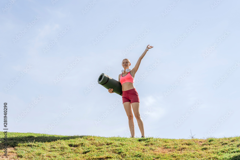 Happy lady holding hand up on green hill with mat Happy lady holding hand up on green hill with mat