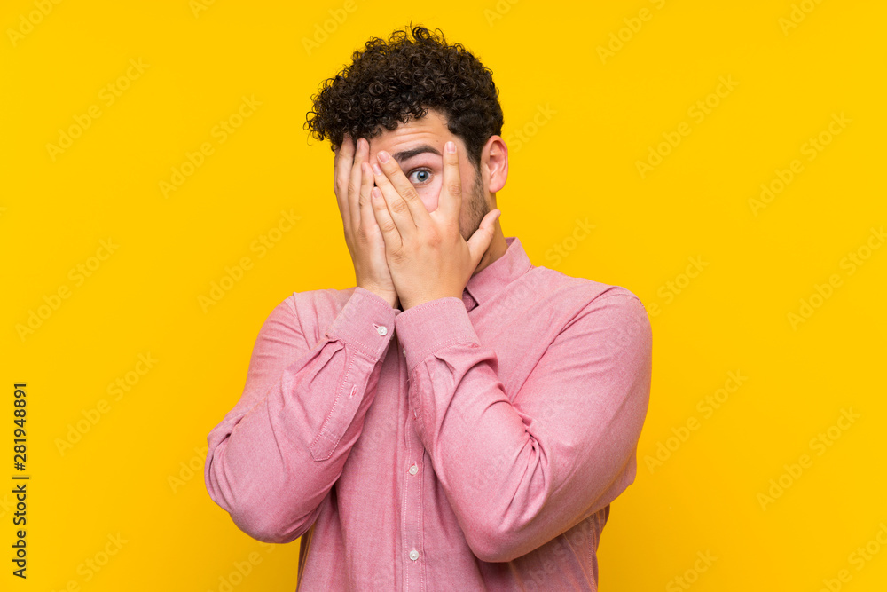 Man with curly hair over isolated yellow wall covering eyes and looking through fingers Man with curly hair over isolated yellow wall covering eyes and looking through fingers