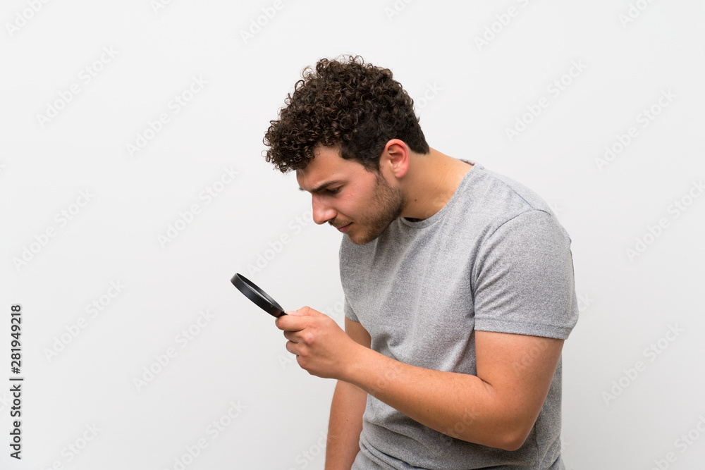Man with curly hair over isolated wall holding a magnifying glass
