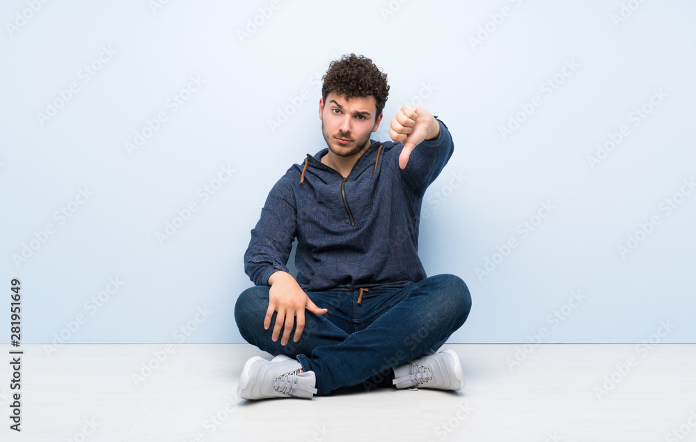 Young man sitting on the floor showing thumb down with negative expression Young man sitting on the floor showing thumb down with negative expression