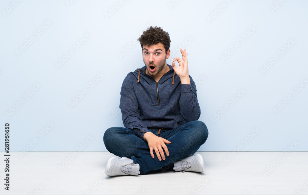 Young man sitting on the floor surprised and showing ok sign Young man sitting on the floor surprised and showing ok sign